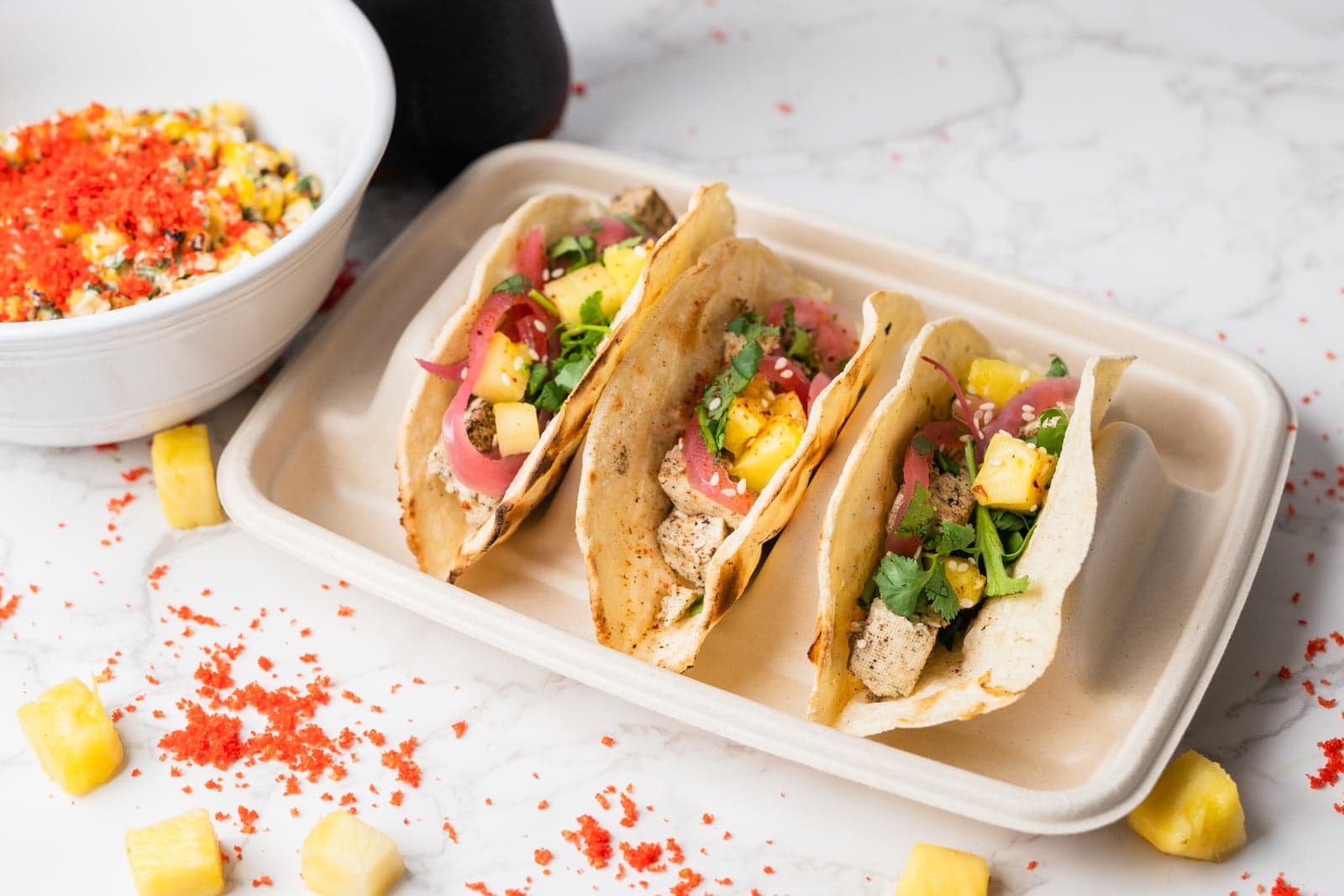 Three Wok-O Taco street tacos with pickled onion, pineapple, and cilantro on a tray beside a bowl of street corn