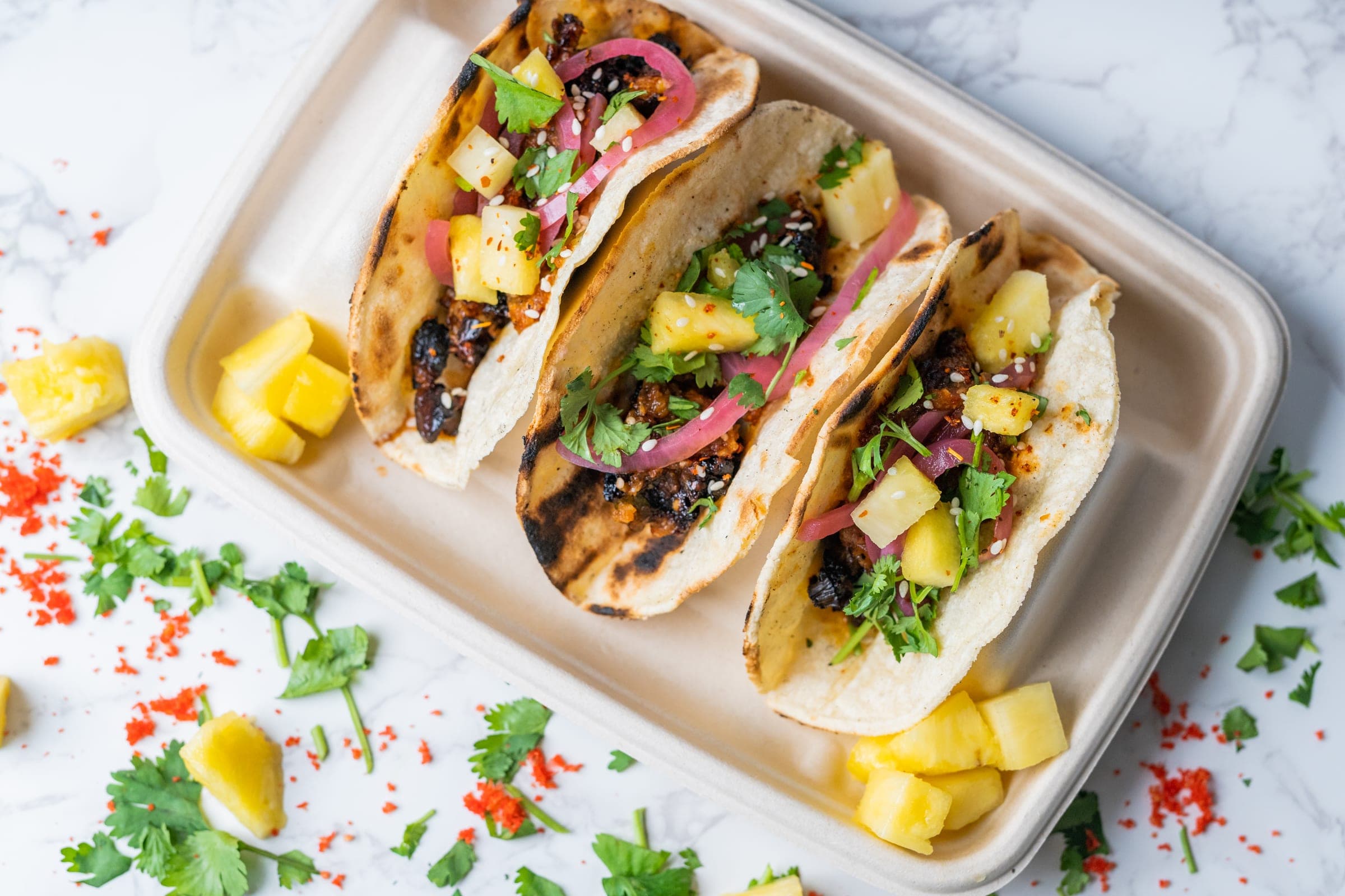 Three pork street tacos topped with pineapple, cilantro, pickled onion, and sesame on a compostable tray