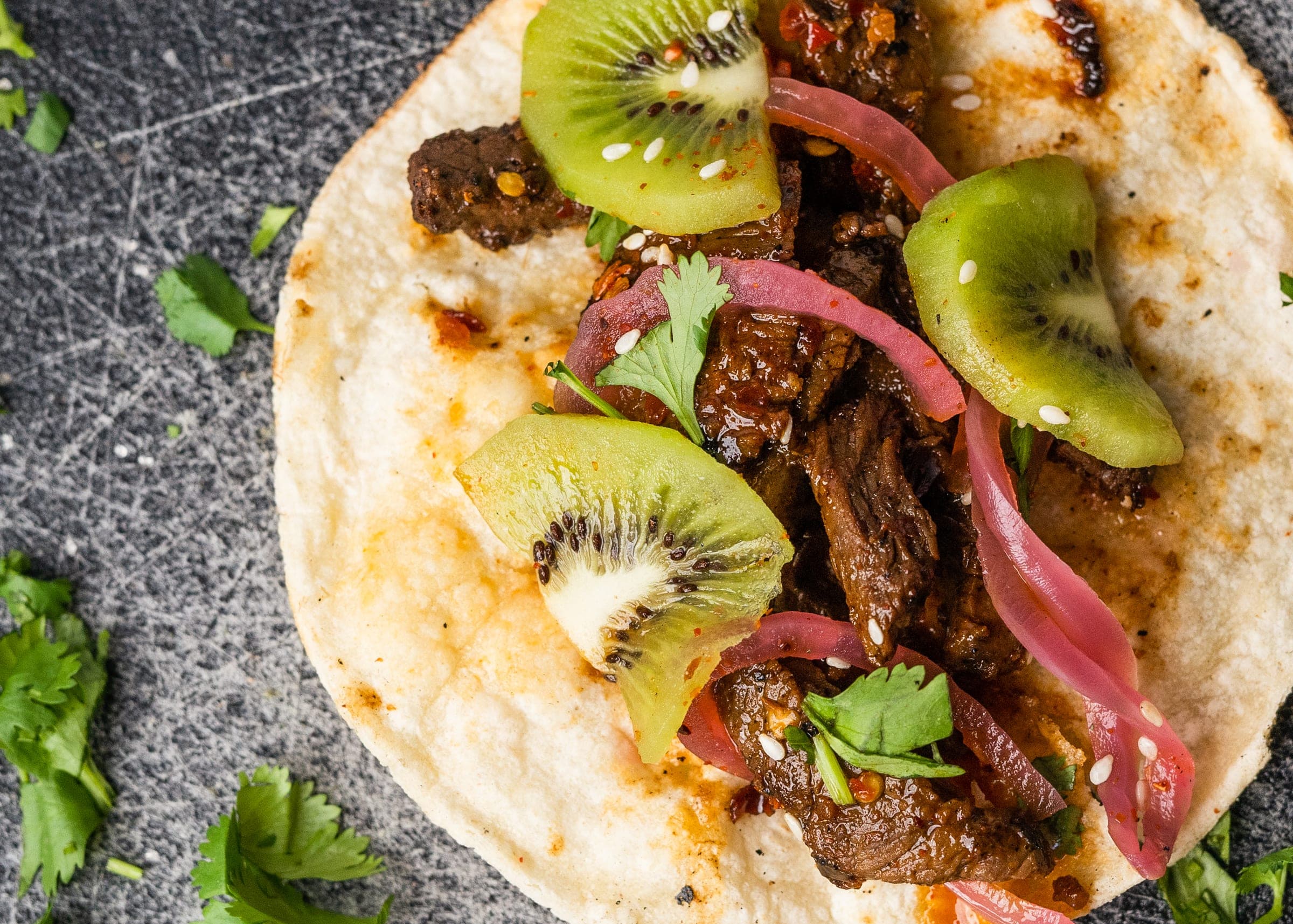 Closeup of a steak street taco with kiwi slices, pickled red onion, cilantro, and sesame seeds
