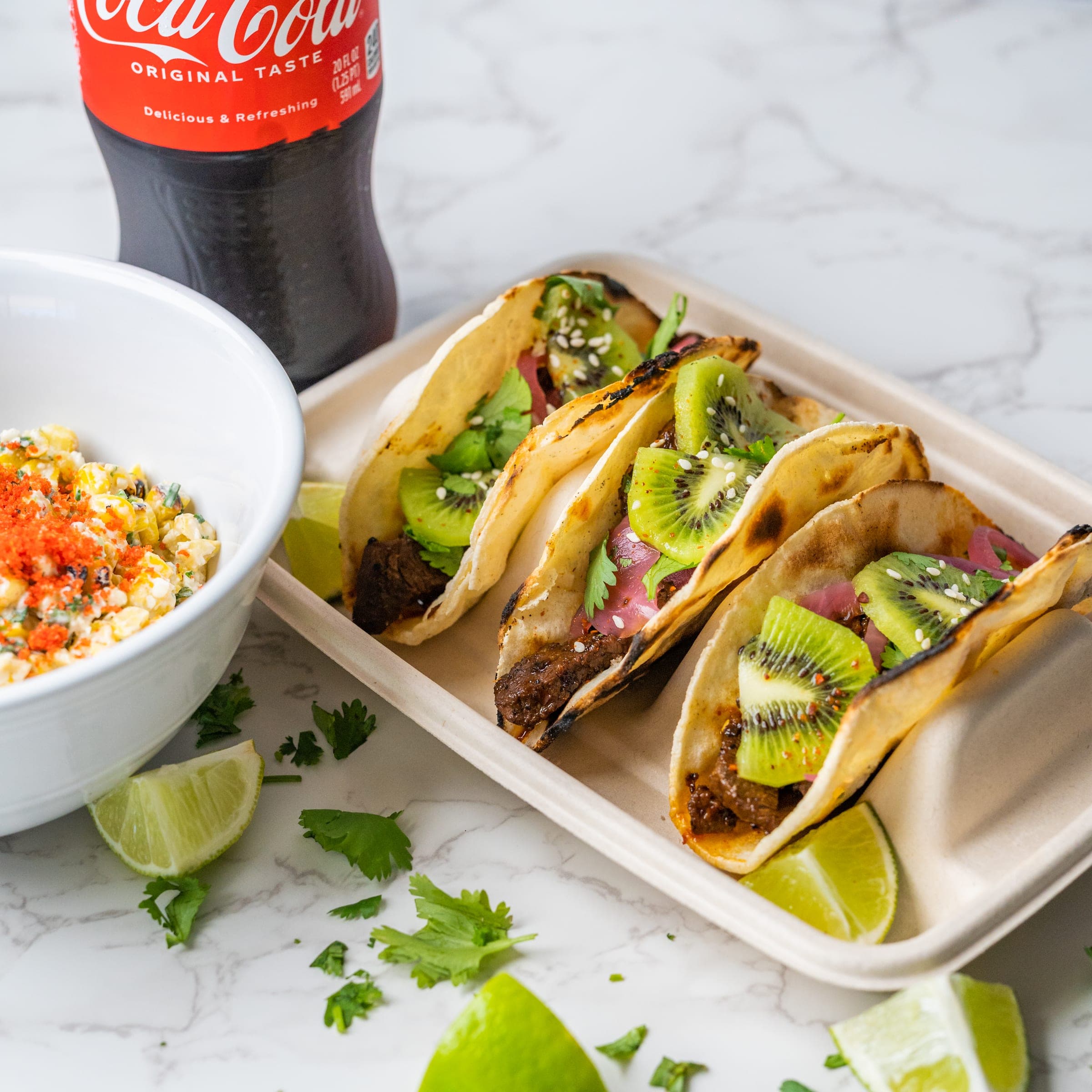 Three steak street tacos with kiwi and pickled onion in a serving tray, beside a bowl of street corn and a bottle of Mexican Coke