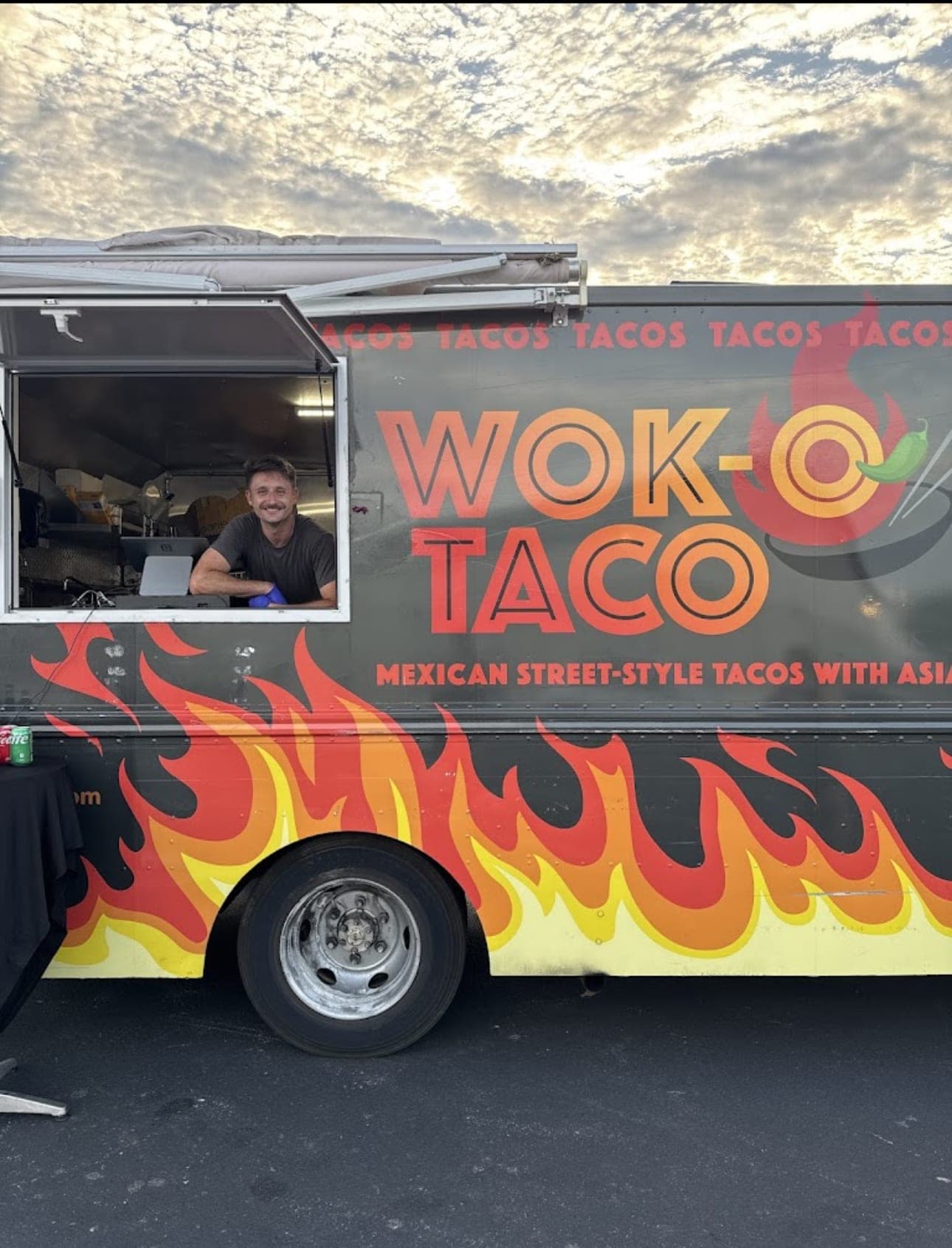 A smiling crew member leaning out the Wok-O Taco truck's service window under an evening sky