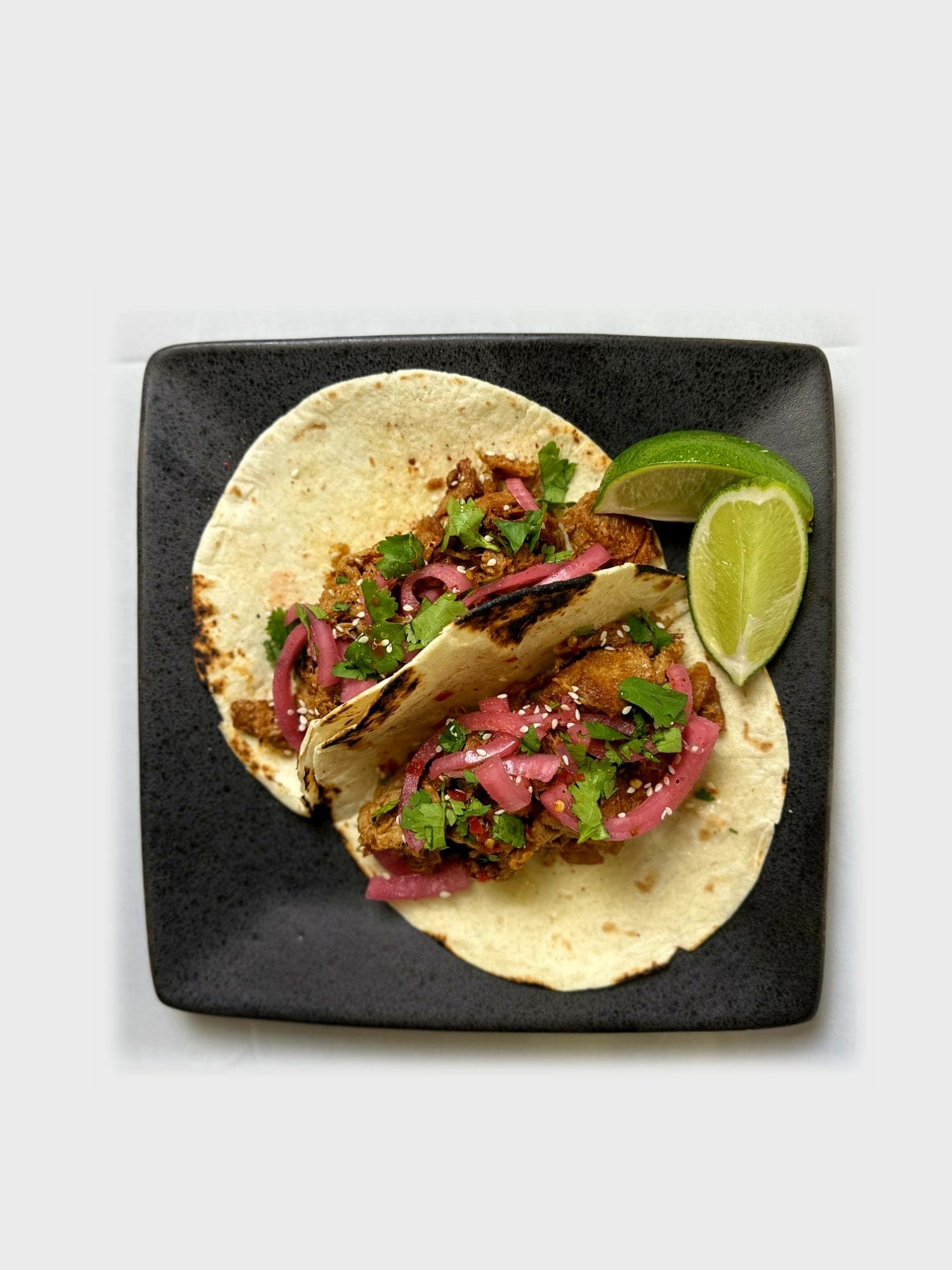 Two street tacos with pickled red onion, cilantro, and sesame seeds beside lime wedges on a square black plate