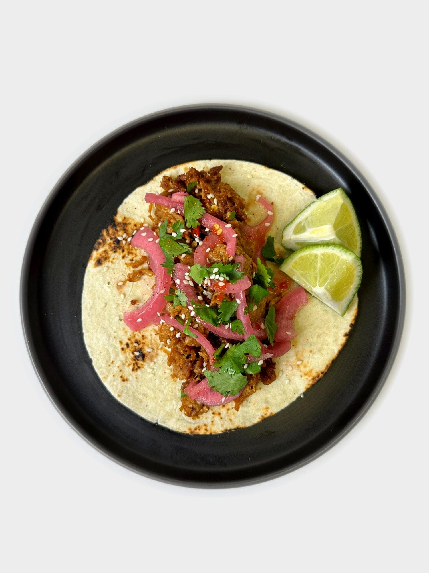 A street taco topped with pickled red onion, cilantro, and sesame seeds, with lime wedges on a black plate