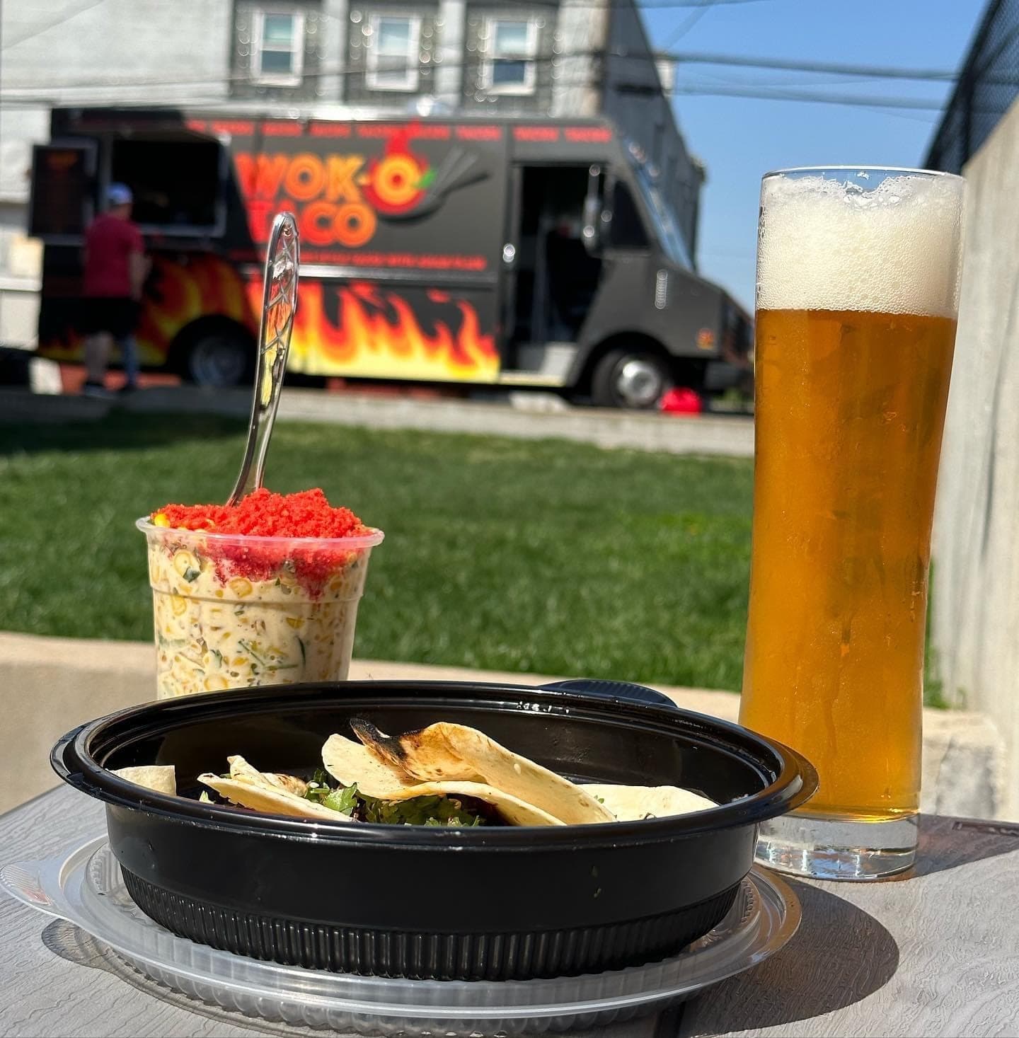 Street tacos in a to-go bowl beside a cup of street corn and a cold drink, with the taco truck in the background